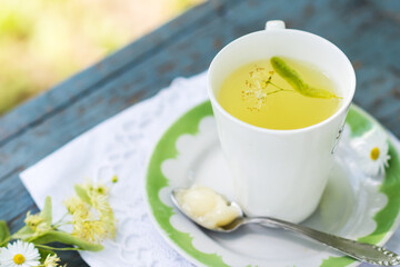 Linden flowers tea in a white cup on a white with green ornament plate with spoon with honey on a blue wooden background and white napkin outdoors, yellow drink of linden flower on  vintage background