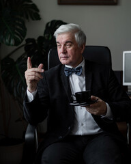 Confident elderly man in a suit and bow tie sits in a chair at his desk and drinks coffee. Portrait of a gray-haired businessman in an office.