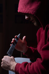 A dangerous bearded man in a red hood holds a sharp knife near his face. A criminal or a killer with knives in the dark.