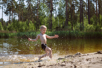 a blond boy in black swimming trunks laughs and runs along the sand on the lake, under his feet spray of water. In the background the forest.
lmage with selective focus