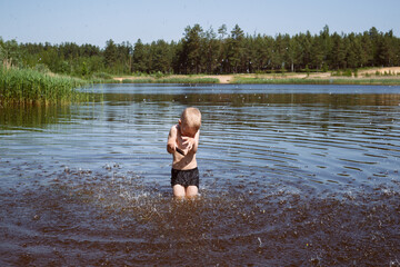 a little blond boy in black swimming trunks on a lake stands in the water and hides from the spray of water. Coniferous forest in the background.
lmage with selective focus