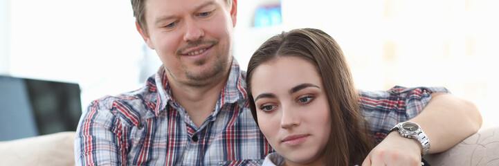 Portrait of pretty couple spending time together reading book on sofa in living room. Smiling boyfriend embracing attractive girlfriend. Love affection and spare time concept