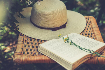 Wildflowers on open book and womens summer hat on old wicker chair in garden, retro filtre, cottage...