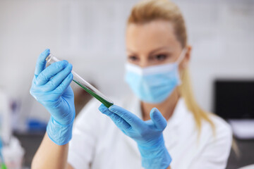 Closeup of lab assistant wit protective gloves and mask on holding test tube with green liquid.