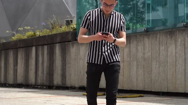 Hombre joven caucasico camina por la calle, mientras revisa las redes sociales en su celular en una ma&ntilde;ana soleada. 