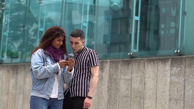 Jovenes amigos en la calle mirando el celular, mientras revisan las redes sociales. Hombre caucasico y chica morena buscan en el celular actividades para realizar. 