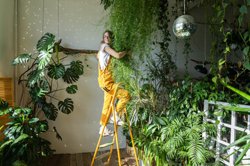 Joyful young woman gardener in orange overalls standing on a stepladder, embracing lush asparagus...