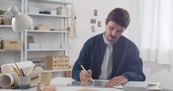 Concentrated man with moustache drawing illustration on tablet . Young guy illustrator using digital notepad and stylus while sitting at table in cosy workplace. Concept of creativity