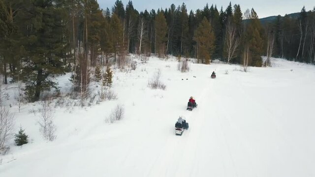 Aerial view of row of snow mobiles ride on a snowy trail near the forest. Clip. Concept of winter extreme sport, people riding snow mobiles along the trees under the heavy sky.