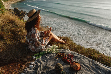 ..Back view young curly hair woman in straw hat  sit on the edge of cliff with ocean view and drink water from stylish eco botle, decorated with macrame, enjoy picnic near the sea