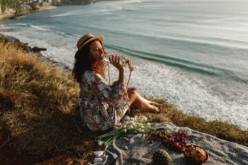 Young curly hair woman in straw hat  sit on the edge of cliff with ocean view and drink water from stylish eco botle, decorated with macrame, enjoy picnic near the sea