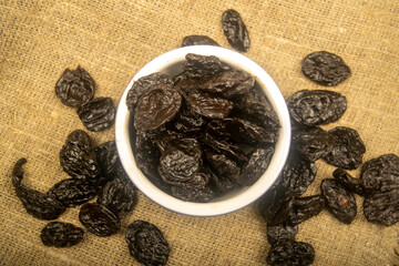 Dried prunes in a metal bowl and dried prunes scattered on a background of coarse-textured burlap....