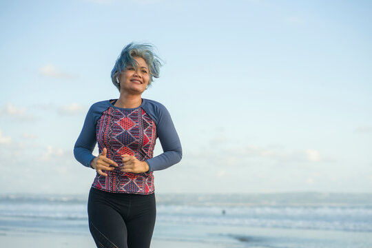 Fit And Happy Middle Aged Woman Running On The Beach - 40s Or 50s Attractive Mature Lady With Grey Hair Doing Jogging Workout Enjoying Fitness And Healthy Lifestyle