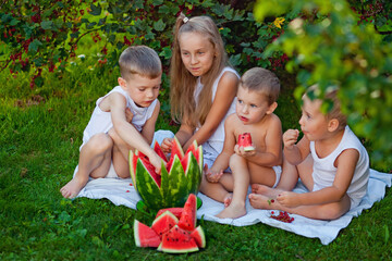 Four children in white in nature sit on the grass. Funny children eat watermelon outdoors in a summer Park. Baby, infant, healthy food.