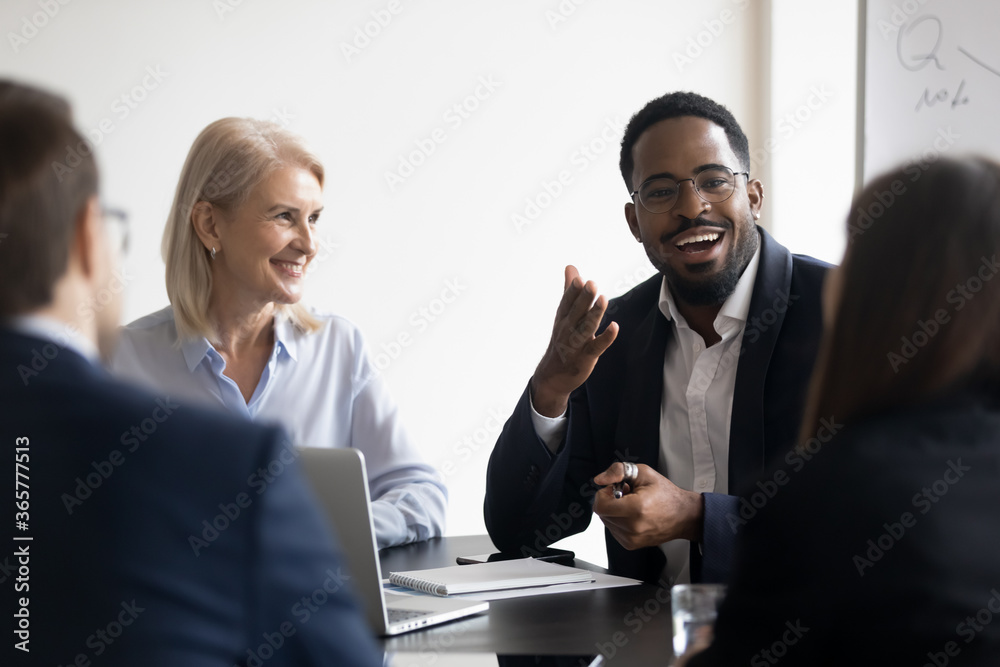 Wall mural Young adults employees gather for briefing, during formal meeting multi ethnic people negotiating listening African skilled trainer participating at corporate workshop sit at desk in modern board room