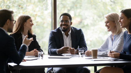 Five positive multi ethnic company staff gather together sit at boardroom desk talking planning...