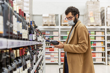 A young man in a supermarket chooses alcohol in the wine section. A brunette in a beige coat with a...