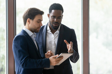 Two multicultural male colleagues businessmen standing in modern office hall using device holding tablet electronic gadget discuss new project, search information, prepare presentation, work together