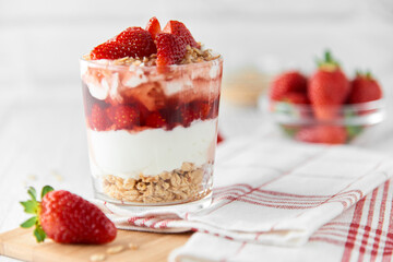 Homemade layered dessert with fresh strawberries, cream cheese or yogurt, granola and strawberry jam in glass on white wood background. Healthy organic breakfast or snack concept. Selective focus.