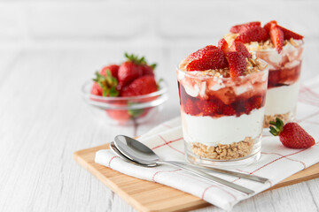 Homemade layered dessert with fresh strawberries, cream cheese or yogurt, granola and strawberry jam in glasses on white wood background. Healthy organic breakfast or snack concept. Selective focus.