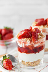 Homemade layered dessert with fresh strawberries, cream cheese or yogurt, granola and strawberry jam in glasses on white wood background. Healthy organic breakfast or snack concept. Selective focus.