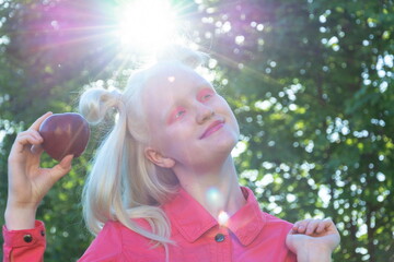 An albino girl in a red blouse against a background of green leaves and sunlight. she holds a red...