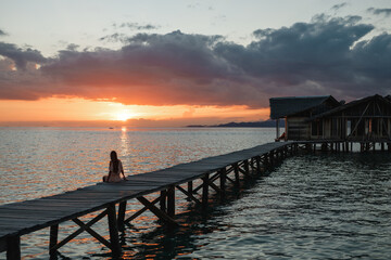 Girl in  dress sitting on a pier on the beach and looking at the water and sunset. Meditation and relaxation.
