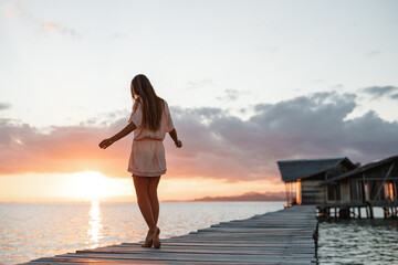 Caucasian woman walks along beautiful seashore on wooden pier during sunset. Young woman in summer dress standing on the pier with peaceful seascape enjoy view on sunset