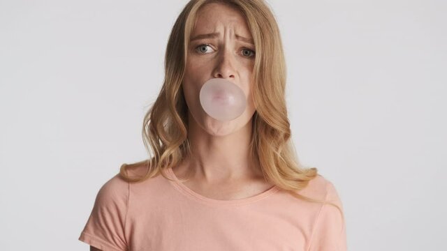 Young funny blond woman scaredly blowing bubblegum over gray background. Candy bubbles