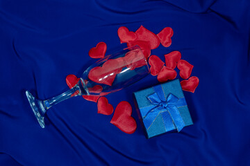Gift box with glass. Crystal glass poured red hearts on blue background.