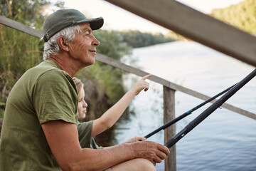 Obraz premium Senior man with his grandson sitting on wooden pontoon with fishing rods in hands, enjoying beautiful nature, little boy pointing at something with his finger.