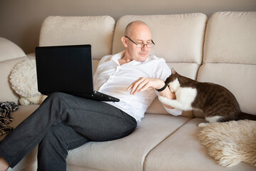 mature man using laptop and playing with cute cat on sofa at home during quarantine of coronavirus covid-19