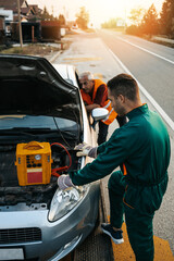 Two road assistant workers in towing service trying to start car engine with jump starter and...