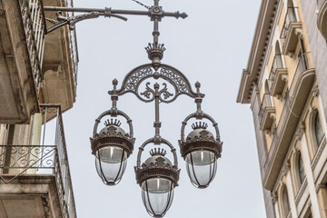 Fototapeta na wymiar Hanging street lamp with three lamps in a black wrought iron design between buildings with balconies in a historic part of the city, gray sky on a cloudy day in Barcelona, ​​Spain