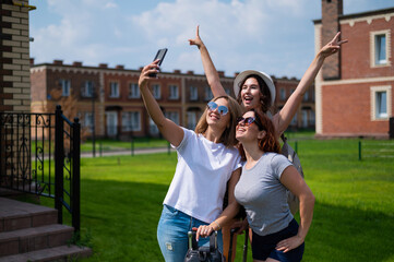 Obraz premium Three happy girls friends go on a trip and take a selfie on a mobile phone. The blonde brunette and the redhead are smiling and photographed on a smartphone in the countryside.