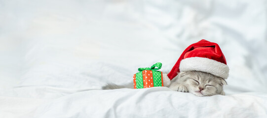 Gray kitten wearing red santa's hat sleeps with gift box on w bed. Empty space for text
