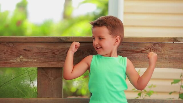 The little cheerful boy in a green t-shirt is showing bicep muscles outdoors
