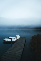 Obraz premium Boats on the lake near jetty on a misty morning. Moody scenery with boats moored in the dock with foggy forest in the background