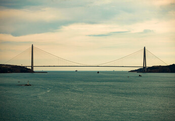 The shot of a third bridge in Istanbul bosporus with couple of ships passing under. Over the Yavuz Sultan Selim Bridge, there are lots of white clouds. Lighthouse in the middle of Marmara Sea.