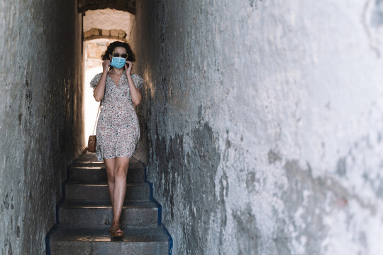 Beautiful curly brunette tourist with a surgical face mask during Covid-19 walking through the narrow and famous street of Correlo de la rectoria in Sitges, Barcelona. Safe Travel in the new normal