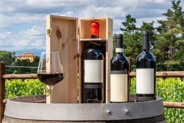 Three bottles including a magnum in the wooden box and a glass of red wine on a wooden barrel with the Tuscan countryside in the background, Italy, on a sunny day - obrazy, fototapety, plakaty