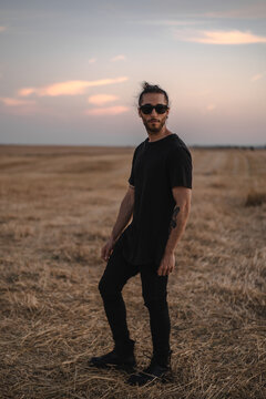 Hombre joven con gafas de sol negras, ropa negra y botas negras, tatuajes y moño paseando por un descampado seco en verano