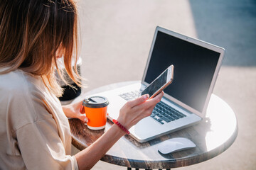 A young girl on a summer playground of a coffee shop works behind a laptop and drinks coffee from a disposable cup 