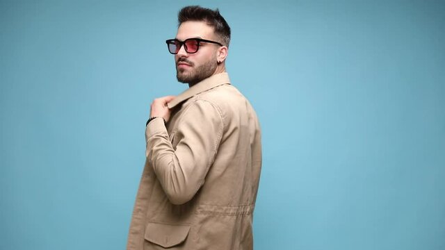 sexy young casual guy wearing sunglasses, adjusting jacket and crossing arms, getting hand through hair and fixing it, holding hand behind neck, touching shoulder and showing off on blue background
