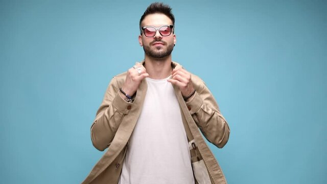 sensual young casual model wearing sunglasses, looking down and smiling, arranging collar and jacket, crossing arms and standing in a fashion pose on blue background