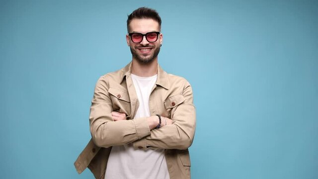 serious young casual man in jacket wearing glasses, starts smiling, crossing arms and standing on blue background