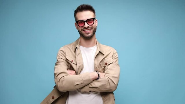 happy young casual guy wearing jacket and sunglasses, smiling and crossing arms, standing on blue background
