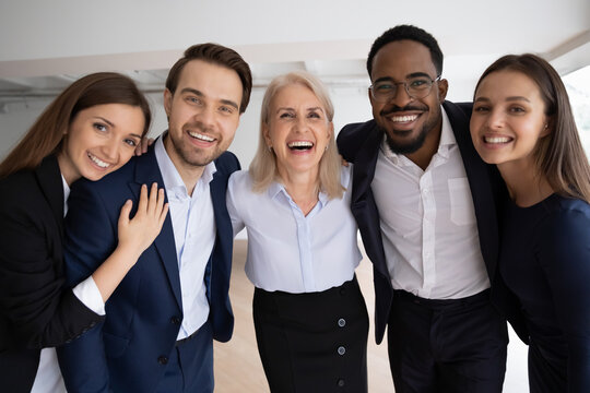 Middle-aged boss company staff members hugging standing together in office laughing looking at camera. Photo shoot corporate album, having gun enjoy friendly relations feels successful and confident