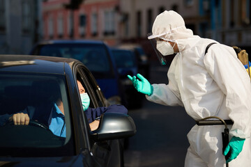 Healthcare worker in protective suit talking to car driver at city checkpoint.