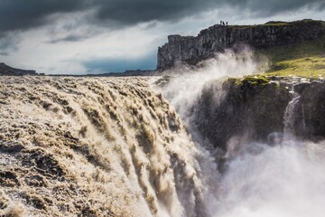 Amazing Dettifoss waterfall  in Vatnajökull National Park in Northeast Iceland. Silhouetts of tourists on the cliff. The falls are 100m wide and have a drop of 44m down to canyon.